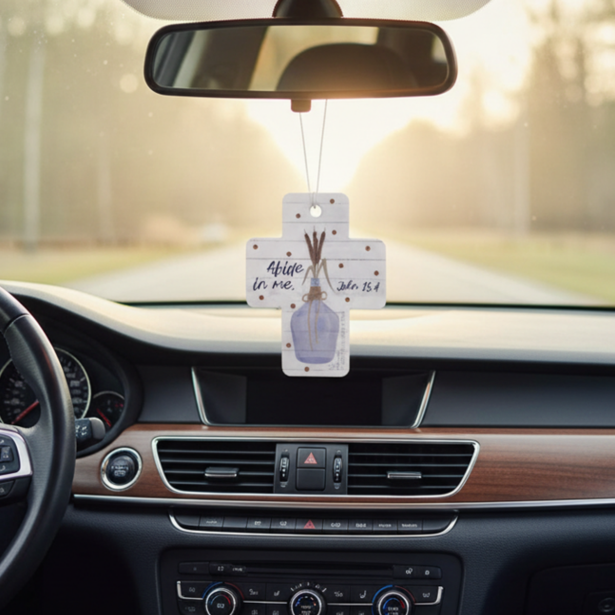 Car air freshener hanging from the rearview mirror with a blurred road background by CTA Christ to All and CTA Gifts.