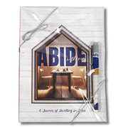 A packaged journal or notebook with a white wood-plank cover, wrapped in clear plastic with a silver string bow. The front features a house-shaped inset photo showing a dining table and chairs, with the large text 'ABIDE with me' and smaller text 'A Journey of Dwelling in You by CTA Christ to All & CTA Gifts.