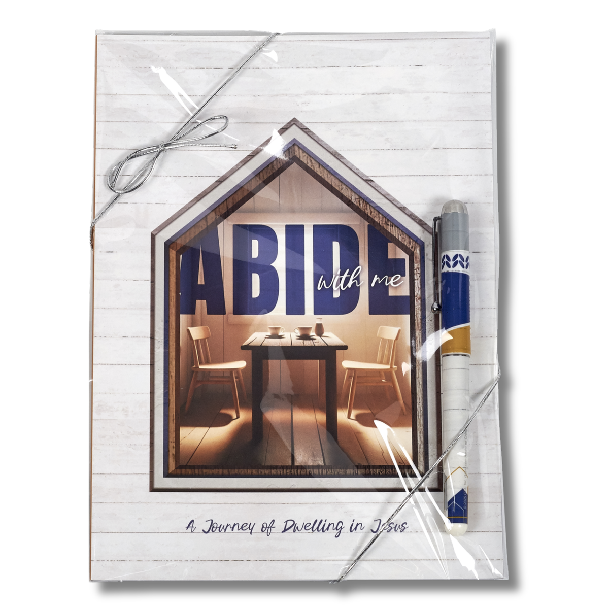 A packaged journal or notebook with a white wood-plank cover, wrapped in clear plastic with a silver string bow. The front features a house-shaped inset photo showing a dining table and chairs, with the large text 'ABIDE with me' and smaller text 'A Journey of Dwelling in You by CTA Christ to All & CTA Gifts.