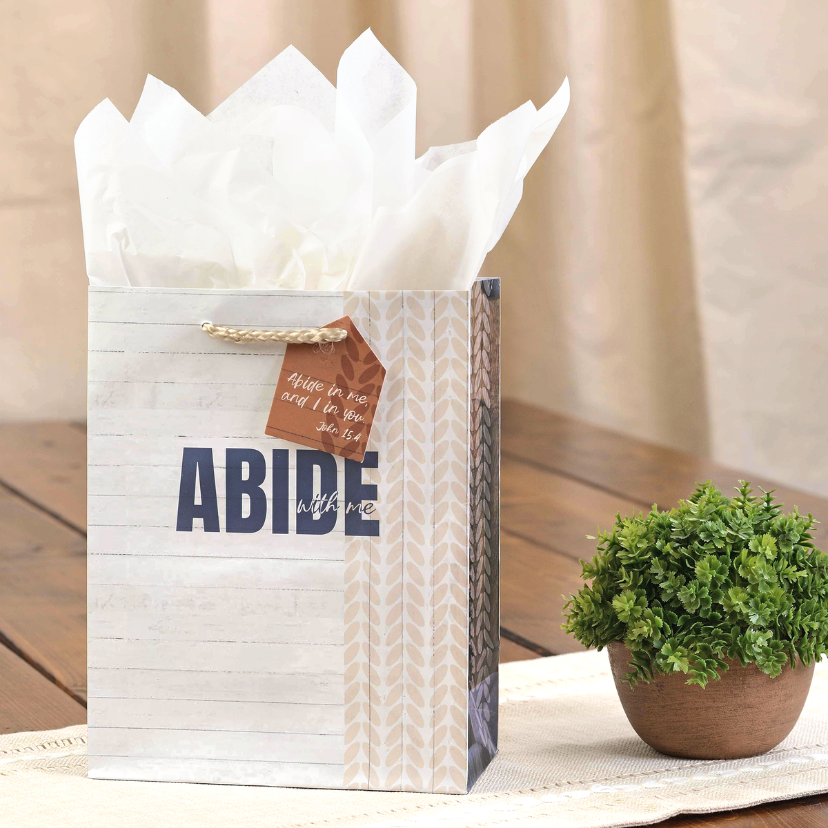A decorative wall art print or canvas showing a rustic white wood plank background with the large text “ABIDE” in dark blue and smaller script text “with me.” Patterned side panels in beige and brown are visible along the left and right edges by CTA Christ to All and CTA Gifts.