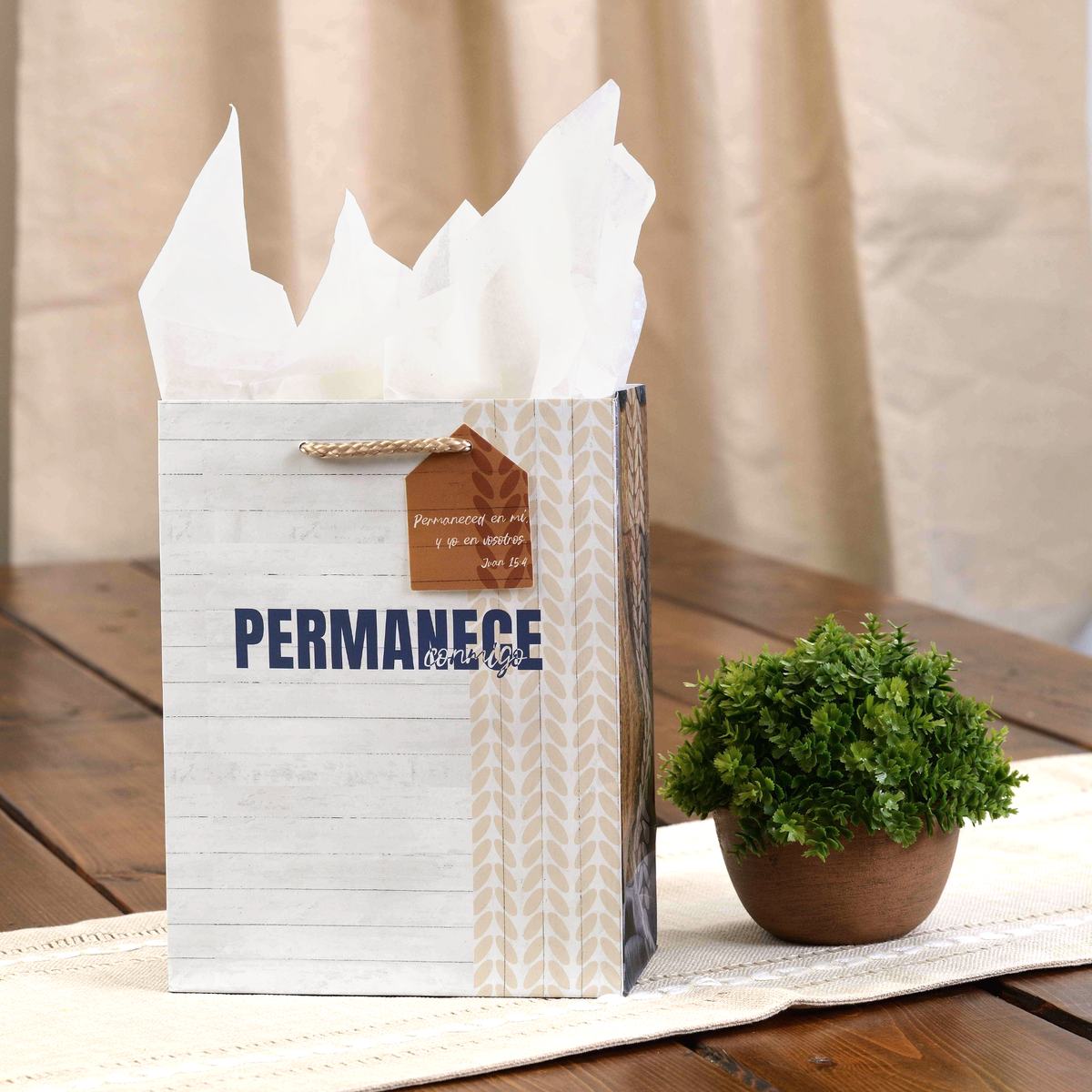 A white and beige gift bag with 'PERMANENCE' printed on the front, filled with white tissue paper and placed on a wooden table next to a small potted green plant by CTA Christ to All and CTA Gifts.