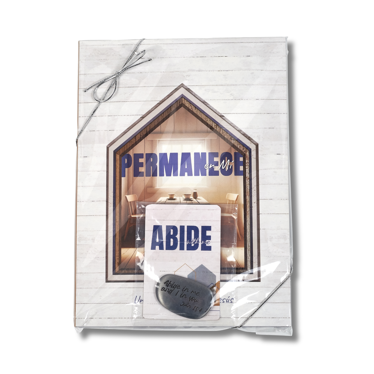 A packaged decorative wall sign or plaque wrapped in clear plastic, featuring a white wood-plank background with a house-shaped window graphic, the words 'PERMANECE' and 'ABIDE' in blue, and a small gray tag attached at the bottom by CTA Christ to All & CTA Gifts.