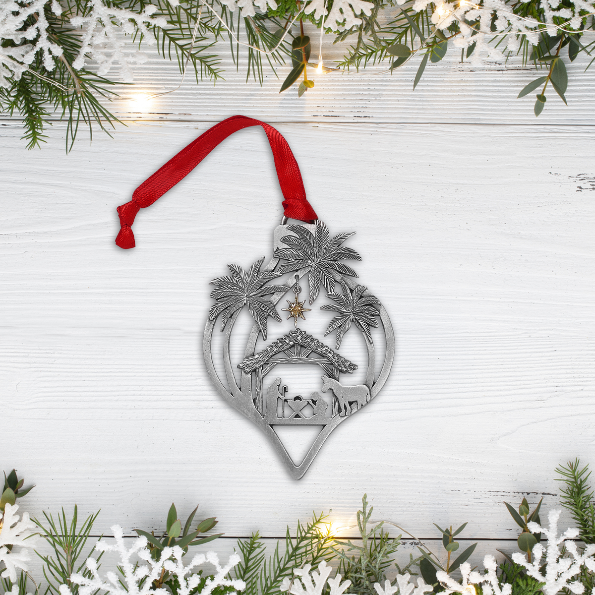 Pewter-finish Nativity ornament with a red hanging ribbon displayed on a white wood surface, surrounded by evergreen branches, snowflake decorations, and warm string lights. The ornament features palm trees, a stable scene with Mary, Joseph, baby Jesus, a donkey, and a gold-tone star by CTA Christ to All and CTA Gifts. Ornaments of Faith® is trademarked.
