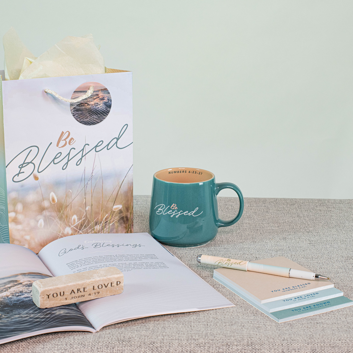 A styled tabletop arrangement featuring a teal ceramic mug with the word 'Blessed' in white script, a matching gift bag, an open book, a pen, and a small notepad on a neutral surface against a pale green wall by CTA Christ to All and CTA Gifts.