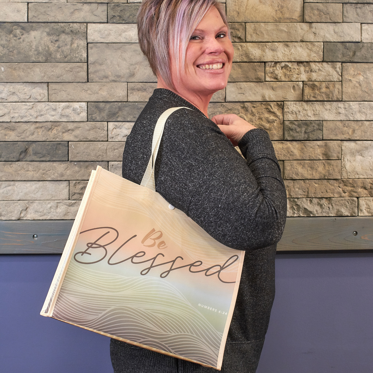 A smiling woman with short pink-tinted hair carries a large canvas tote bag over her shoulder. The tote bag has a cream and beige background with wavy line patterns and the words 'Be Blessed' in elegant script by CTA Christ to All and CTA Gifts.