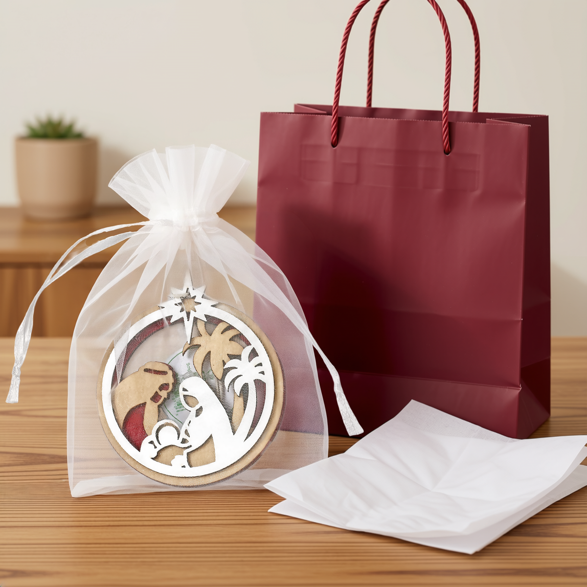 Laser cut wooden Christmas Nativity ornament featuring Mary, Joseph, and Baby Jesus beneath the Star of Bethlehem, packaged in a white organza gift bag with tissue paper and a burgundy gift bag in the background. Decorative Christian Christmas ornament ideal for holiday gifting, church events, stocking stuffers, and faith-based Christmas décor, by CTA Christ to All and CTA Gifts.