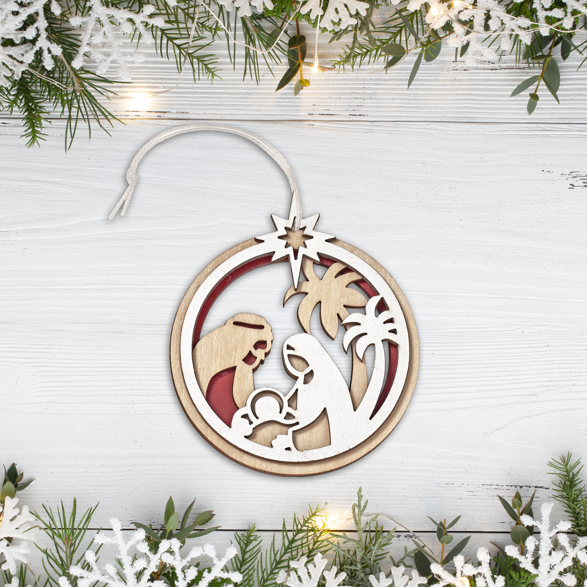 Laser cut wooden Christmas Nativity ornament featuring Mary, Joseph, and Baby Jesus beneath the Star of Bethlehem. String hanger, displayed on a white wood background with festive greenery and lights and faith-based Christmas décor, by CTA Christ to All and CTA Gifts.