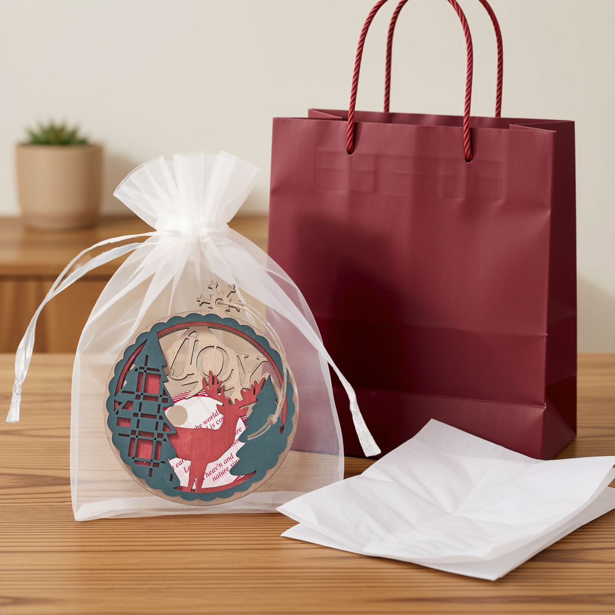 Laser-cut wooden Christmas ornament featuring evergreen trees, deer, the word “JOY”, string cord hanger, packaged in a white organza gift bag with tissue paper and a burgundy gift bag in the background. Decorative Christian Christmas ornament ideal for holiday gifting, church events, stocking stuffers, and faith-based Christmas décor, by CTA Christ to All and CTA Gifts.