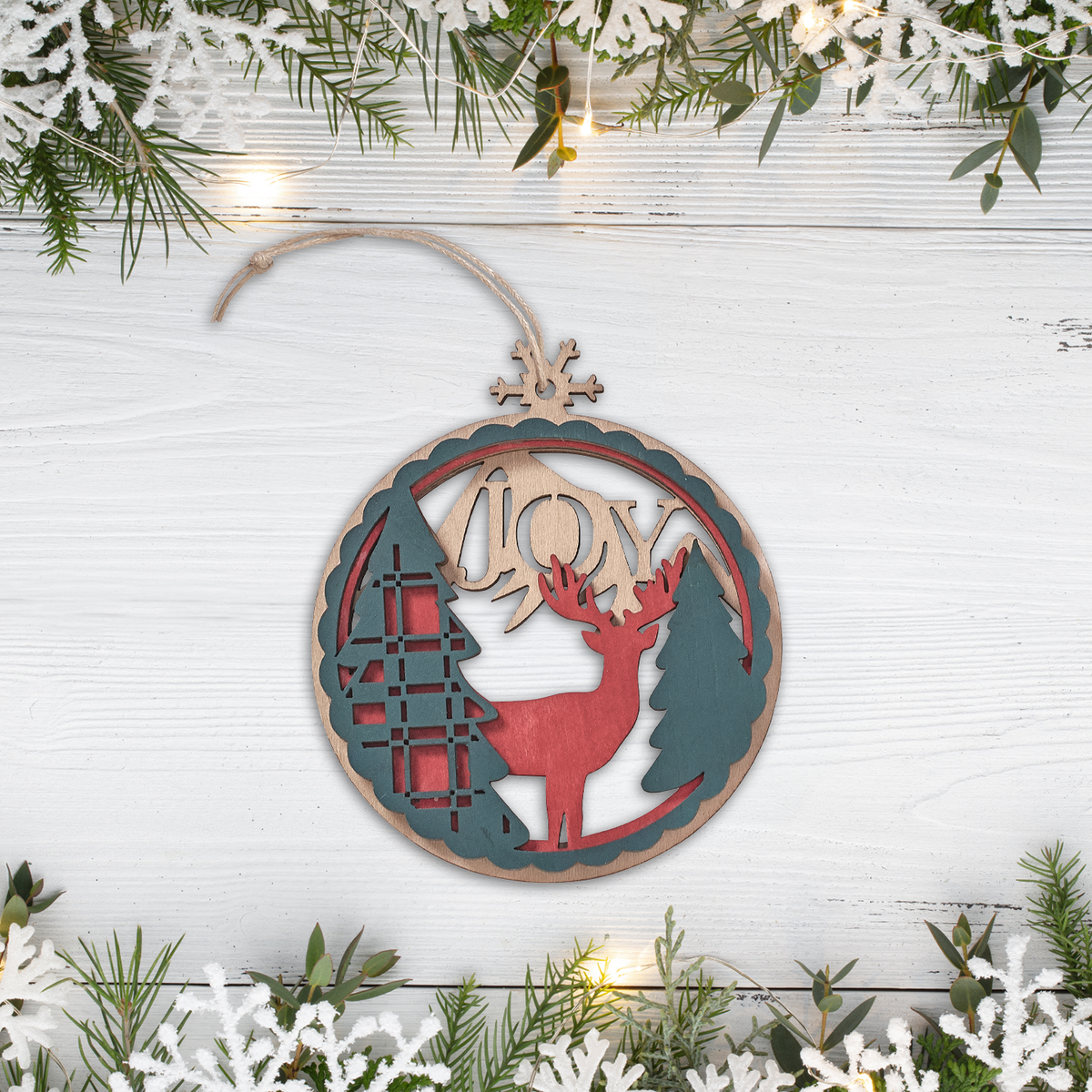 Laser-cut wooden ornament featuring evergreen trees, deer, the word “JOY”, string cord hanger displayed on a white wood background with festive greenery and lights and faith-based Christmas décor, by CTA Christ to All and CTA Gifts.