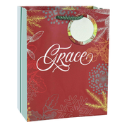 A red gift bag with white script text reading 'Grace' and a circular white insert, decorated with gold and silver winter botanical illustrations including pine branches, snowflakes, and holly leaves. The bag has a green braided handle and a light blue side panel by CTA Christ to All and CTA Gifts.
