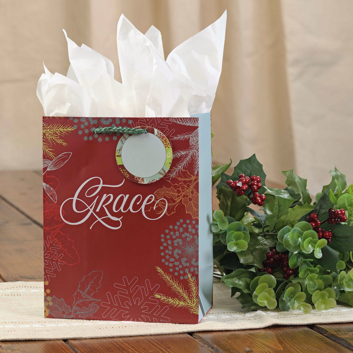 A red Christmas-themed gift bag with white tissue paper inside, featuring the name 'Grace' in white script and gold and silver holiday patterns. The bag is styled on a wooden surface next to green holly leaves and red berries against a neutral beige background by CTA Christ to All and CTA Gifts.