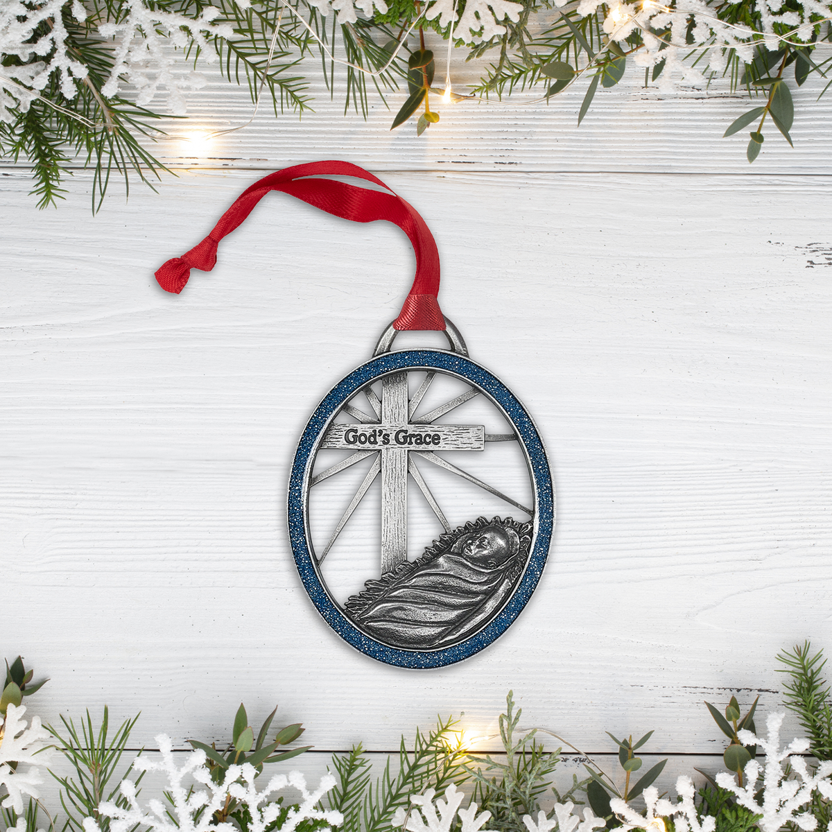 Round Christmas ornament featuring baby Jesus in a manger beneath a silver cross engraved with “God’s Grace,” accented by a blue glitter frame and red hanging ribbon. Ornament is styled on a festive white wood background with greenery, snowflake décor, and twinkling lights by CTA Christ to All and CTA Gifts.