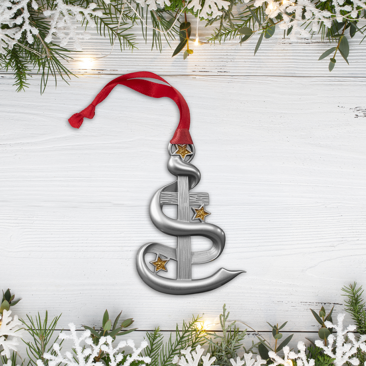 A silver-toned metal Christmas ornament featuring a relief carving of the Holy Family, hanging from a red ribbon on a decorated Christmas tree branch with warm lights in the background.