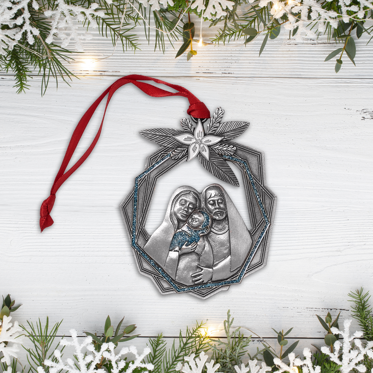 A Christmas ornament shaped like a silver ribbon wrapped with a red ribbon and small gold stars, displayed on a white wood background with festive greenery and lights.