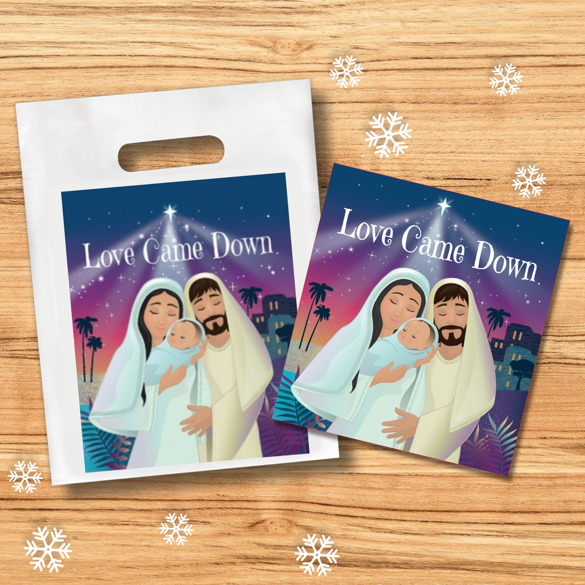 Product display featuring a white handled gift bag and matching “Love Came Down” booklet placed on a light wood background. Both items showcase a colorful Christmas cover illustration with a nativity scene beneath a bright star and the title “Love Came Down.” White snowflake graphics are scattered around the arrangement, creating a festive holiday presentation by CTA Christ to All and CTAKids.