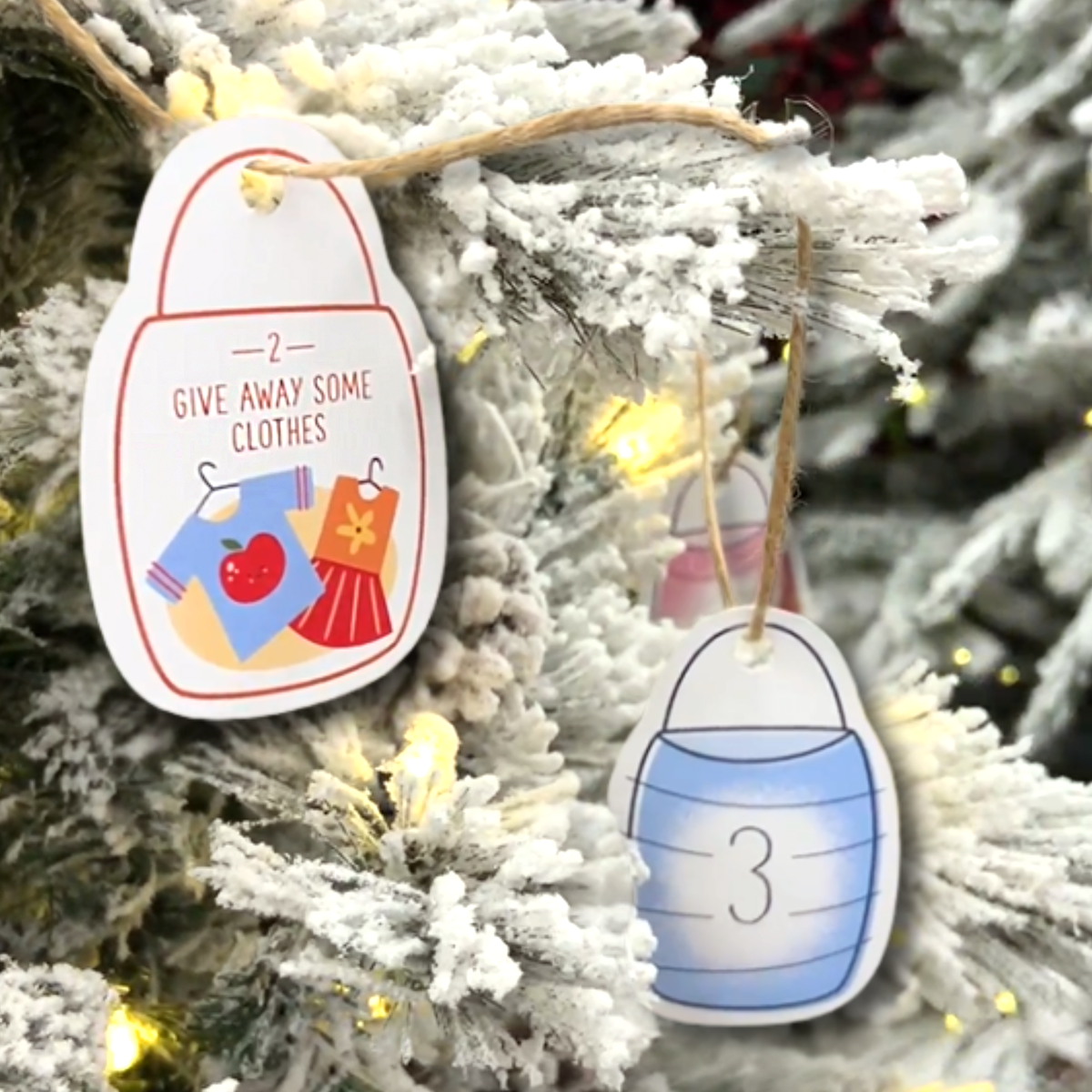 Two Christmas advent-style paper tags hang from a frosted Christmas tree with warm string lights. One tag says “GIVE AWAY SOME CLOTHES” with illustrations of a blue dress and a red skirt, and the other tag shows the number 3 inside a blue hat outline by CTA Christ to All and CTAKids.