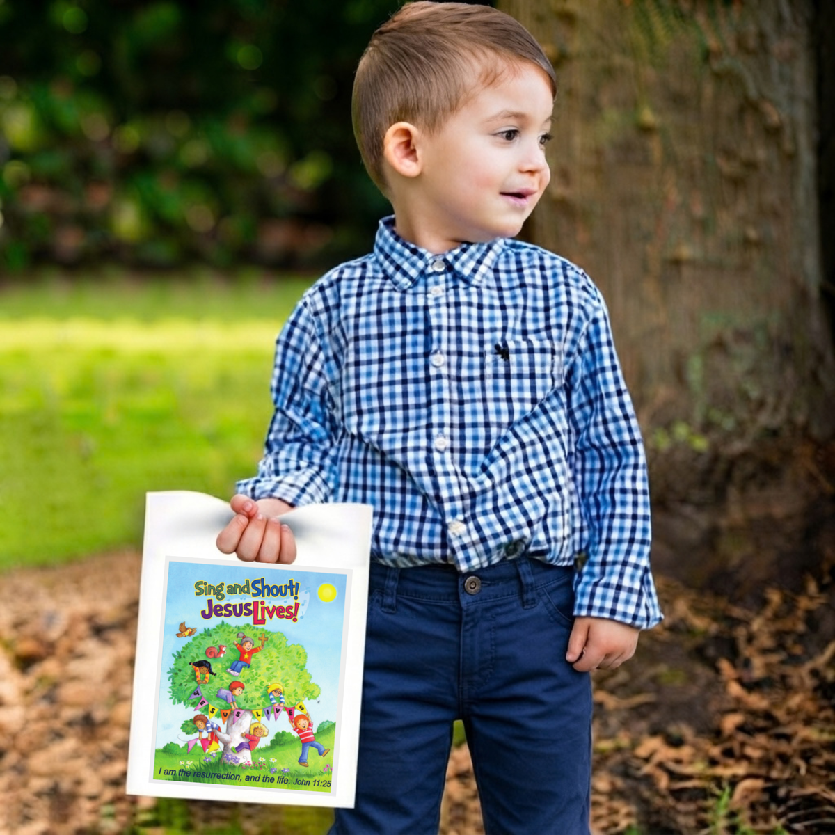 A young boy wearing a blue and white gingham shirt and navy pants stands outdoors in a park-like setting, holding a children's book titled 'Sing and Shout! Jesus Lives!' by CTA Christ to All and CTAKids.