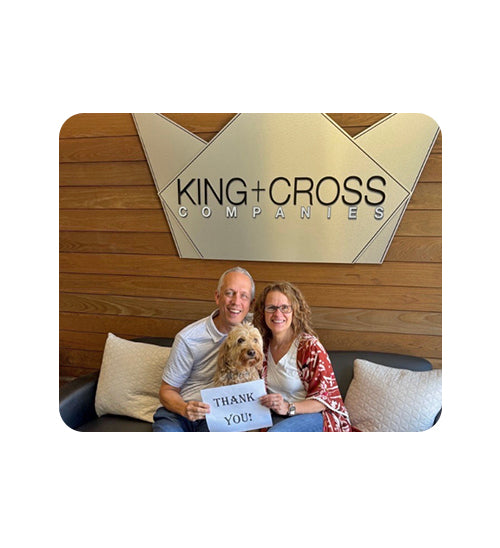 A man and a woman sitting on a couch with a small dog, holding a sign that says 'THANK YOU!' in front of a wooden wall with a large KING+CROSS COMPANIES sign.