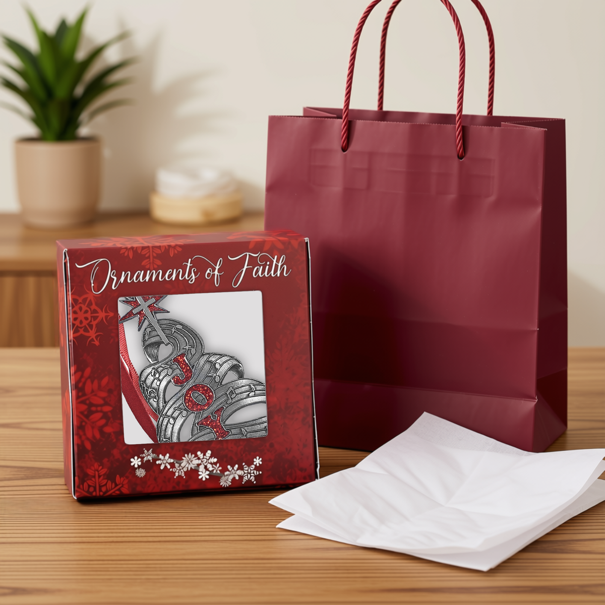 Red "Ornaments of Faith" gift box displaying a pewter-finish Christmas tree ornament made from flowing sheet music with red glitter letters spelling “JOY,” shown alongside a red gift bag and white tissue paper for easy gift giving by CTA Christ to All and CTA Gifts. Ornaments of Faith® is trademarked.