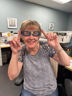 Person sitting at a desk in an office workspace, wearing eclipse viewing glasses decorated with a star design and raising both index fingers, with cubicles and office décor in the background by CTA Christ to All and CTAKids.