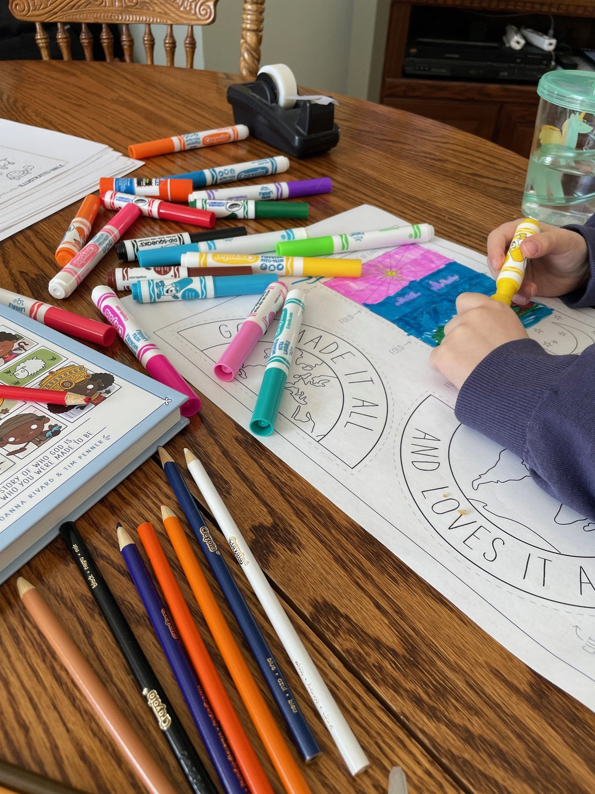Colorful markers and pencils on a table with a child coloring a sheet.