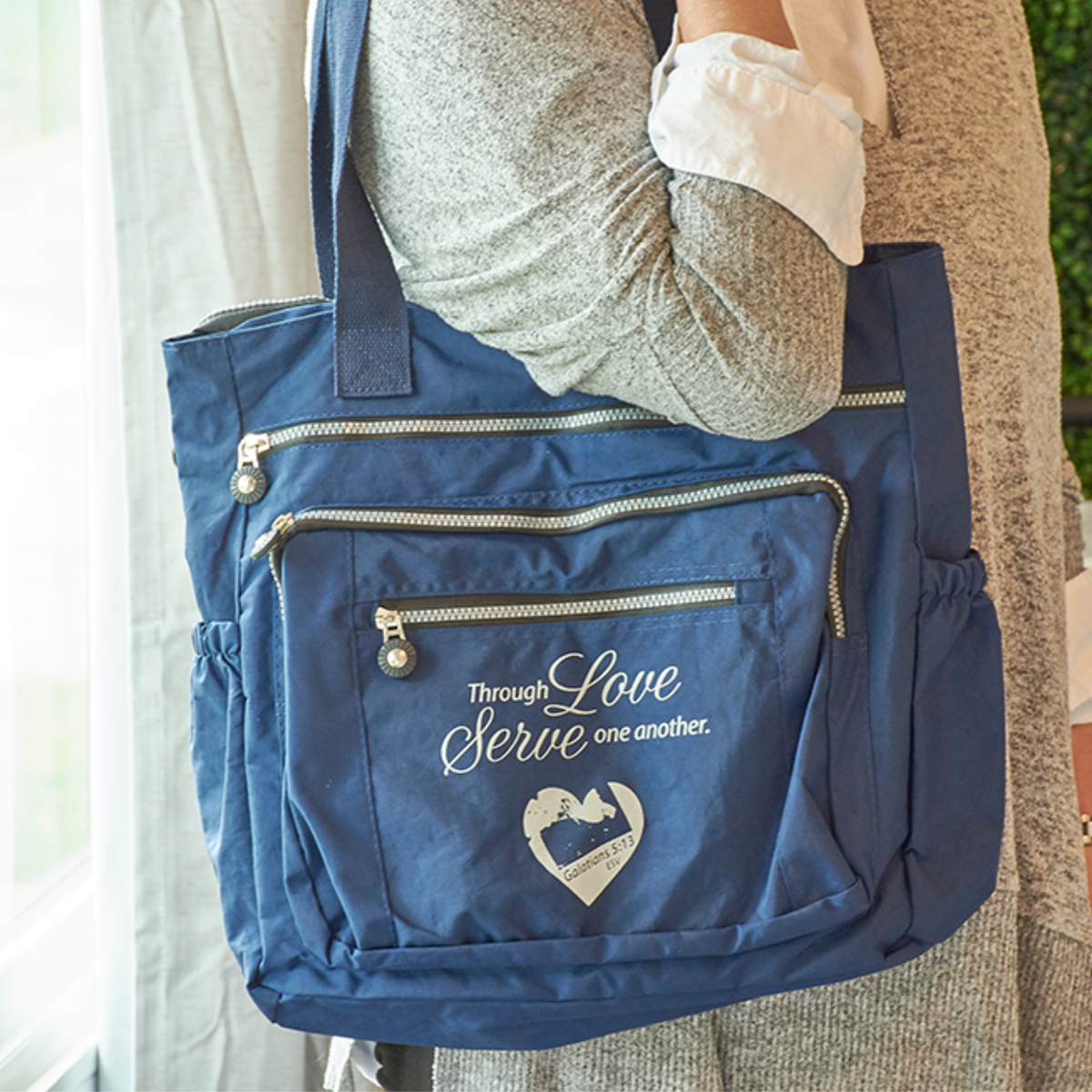 A navy blue fabric tote bag with multiple zippered pockets and white embroidered text reading 'Through Love Serve one another' with a heart design, carried by a person wearing a gray knit top and beige cardigan by CTA Christ to All and CTA Gifts.