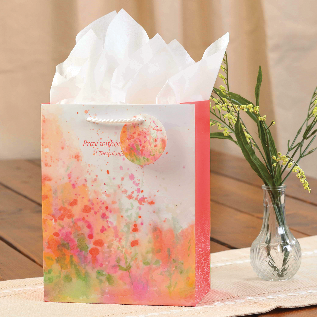 Floral gift bag with white tissue paper, placed on a wooden surface with a vase of flowers in the background by CTA Christ to All and CTA Gifts.