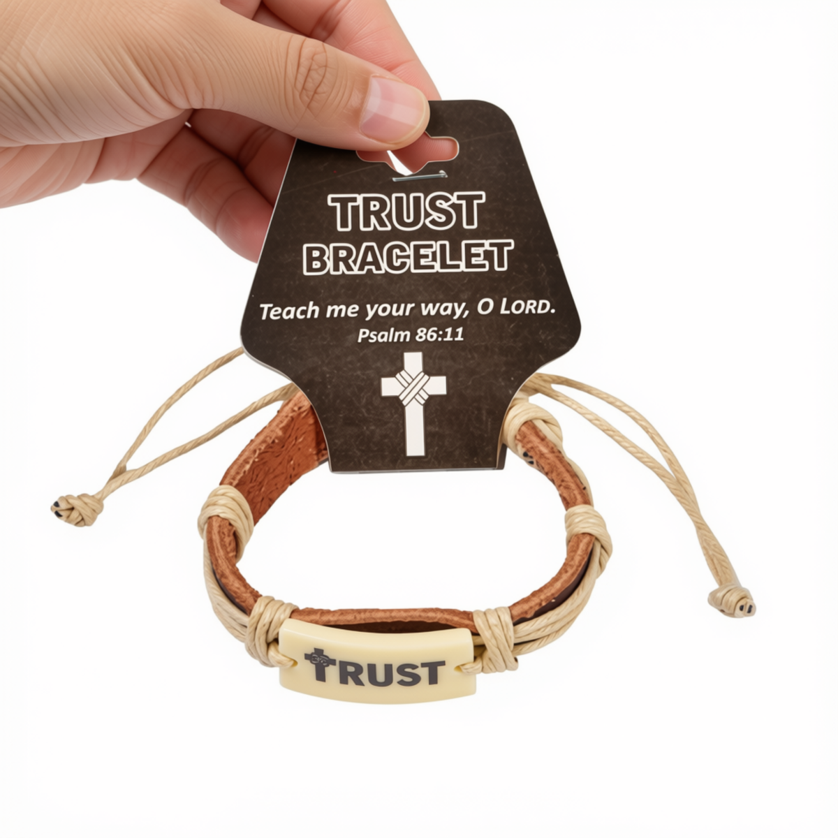 A hand holds a brown cardboard hanger displaying a leather bracelet. The bracelet has tan and brown leather wraps, a beige cord, and a light wood-toned rectangular plate engraved with the word 'TRUST' and a cross by CTA Christ to All & CTAKids.