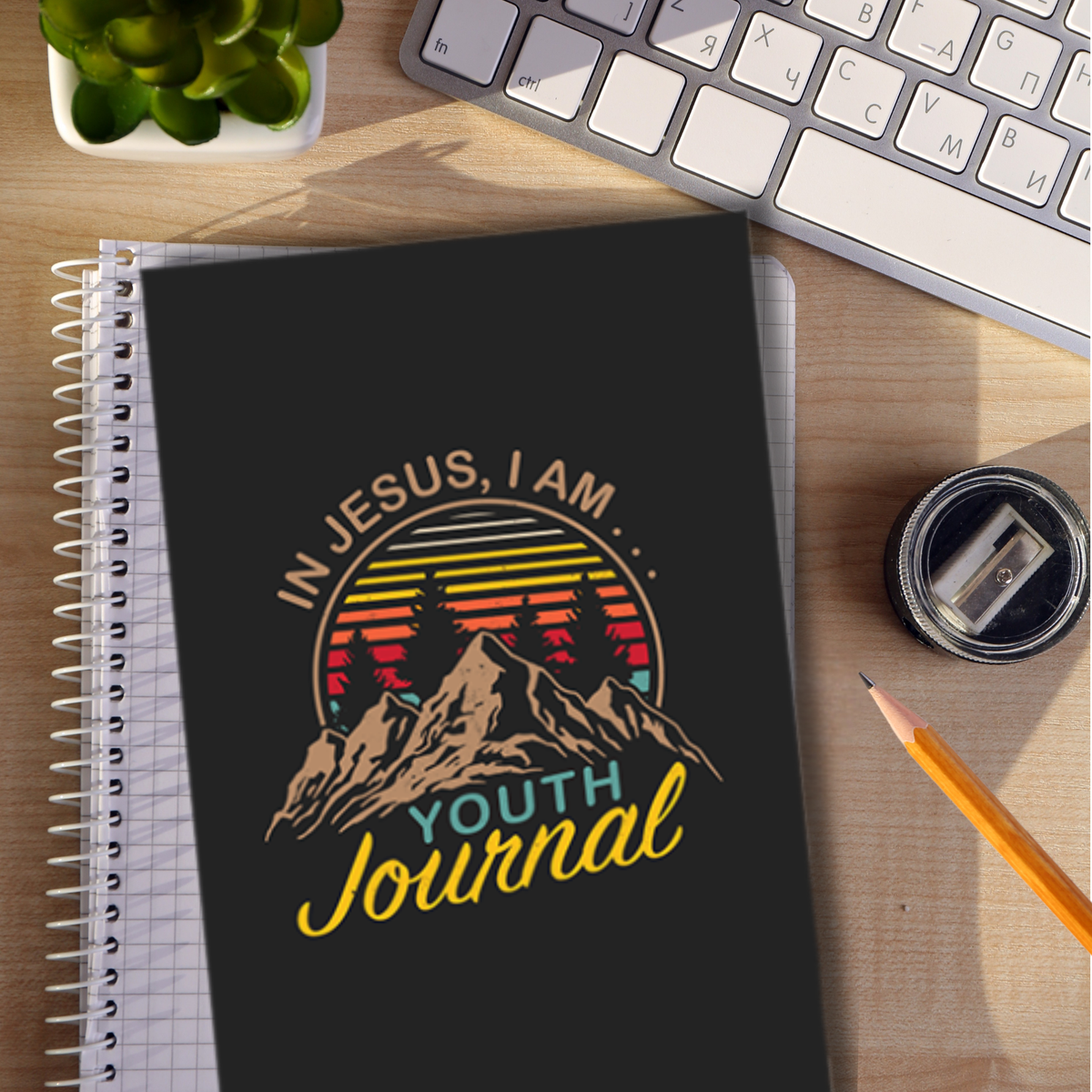 Journal with 'In Jesus, I Am... Youth Journal' design on a desk with a keyboard and stationery items by CTA Christ to All and CTAKids.
